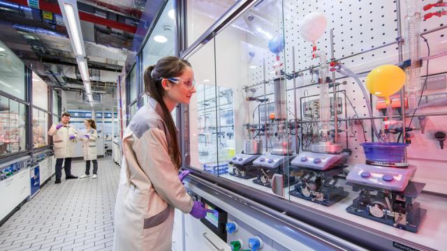 A scientist in a lab coat and safety glasses operates equipment in a laboratory with other scientists working in the background.