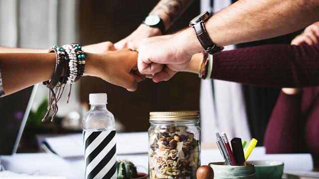 Four people fist-bumping over a table with a water bottle and a jar of snacks.