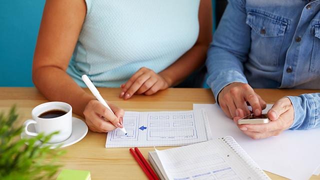 Two people sit at a table with coffee, notebook, and smartphone; one is writing on graph paper while the other holds the phone.