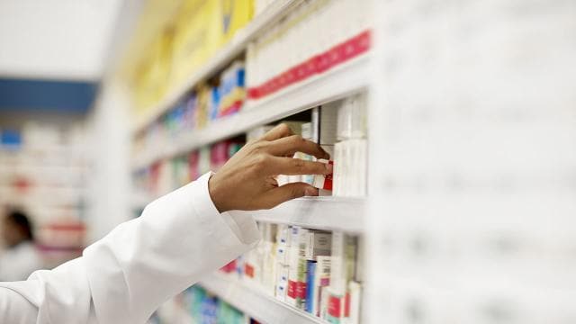 A pharmacist's hand reaches for a medication box on a shelf filled with various pharmaceutical products.