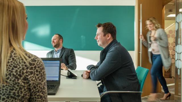 Four people in a meeting room, two men sitting at a table with laptops, a woman entering, and another woman facing them.