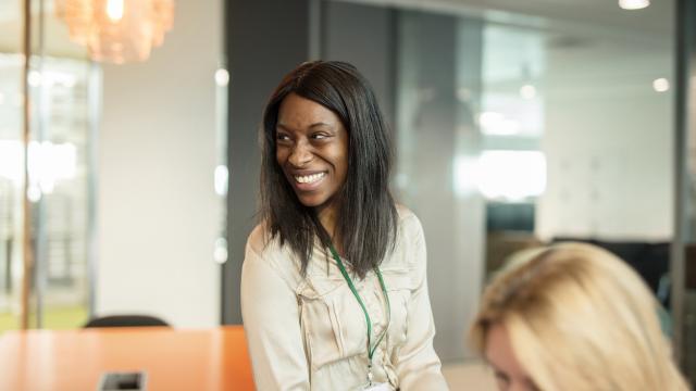 A woman in a beige blouse smiles while standing in a modern office setting.