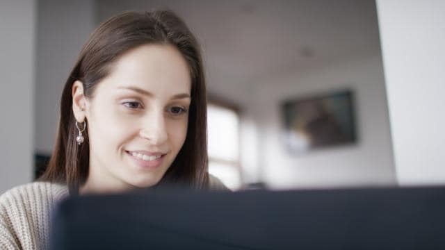 A woman is smiling while looking at a computer screen in a softly lit room.