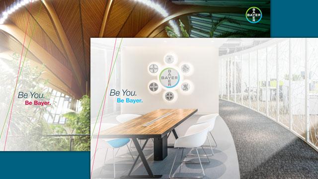 Modern office space with a long wooden table, white chairs, and a wall displaying circular logos. Text reads: "Be You. Be Bayer." Ceiling features wooden beams and skylights.