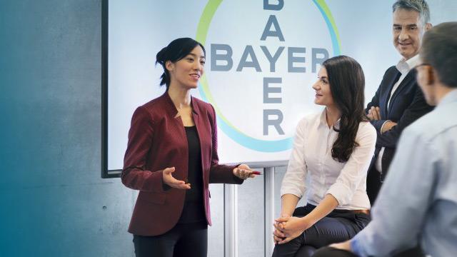A woman presents in front of a screen displaying the Bayer logo, with several colleagues listening attentively.