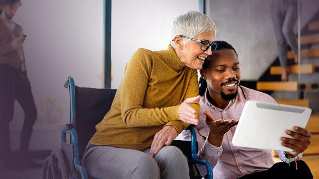A woman in a wheelchair and a man sit together, smiling and looking at a tablet.