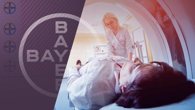 A healthcare professional assists a patient lying on their back entering an MRI scanner. The Bayer logo is visible on the left side of the image.