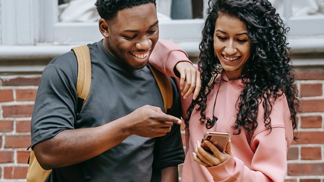 Two people stand outdoors by a brick wall, smiling and looking at a smartphone. One person points at the phone, and both appear engaged.