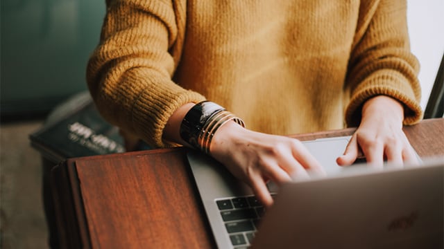 A person in a yellow sweater types on a laptop at a wooden desk.