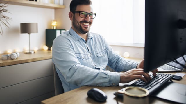 Man wearing glasses, smiling while typing on a keyboard at a desk with a computer monitor.