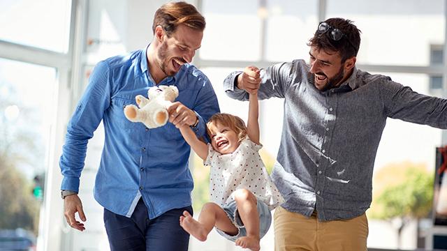 Two adults are joyfully swinging a child between them inside a bright room. The child holds a stuffed toy.