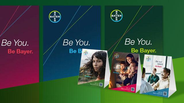 Bayer promotional posters featuring the slogan "Be You. Be Bayer." with images of diverse individuals in various work settings.
