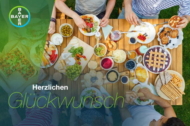 Aerial view of a group of people sharing a meal at a wooden table outdoors with various dishes and snacks, featuring a Bayer logo and "Herzlichen Glückwunsch" text overlay.