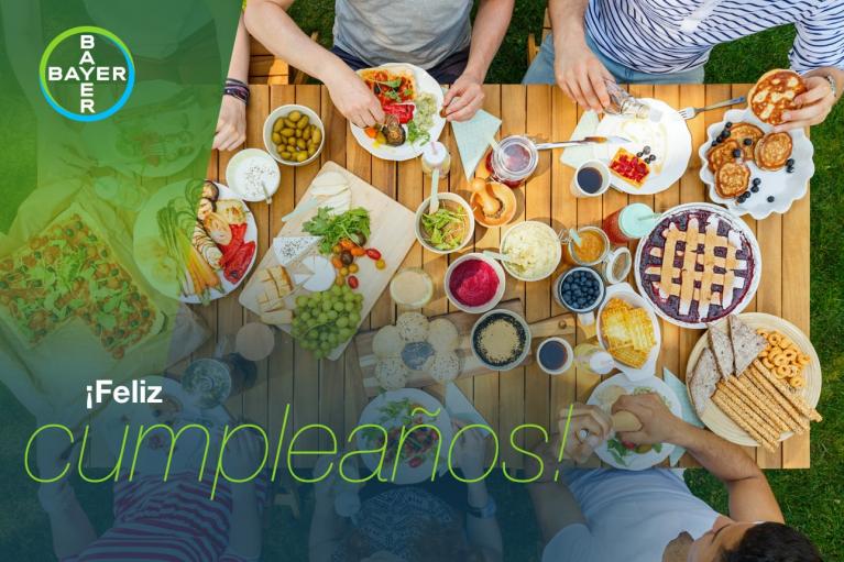 Aerial view of people eating a variety of foods at a wooden table outdoors. The text "¡Feliz cumpleaños!" and a Bayer logo are overlaid on the image.