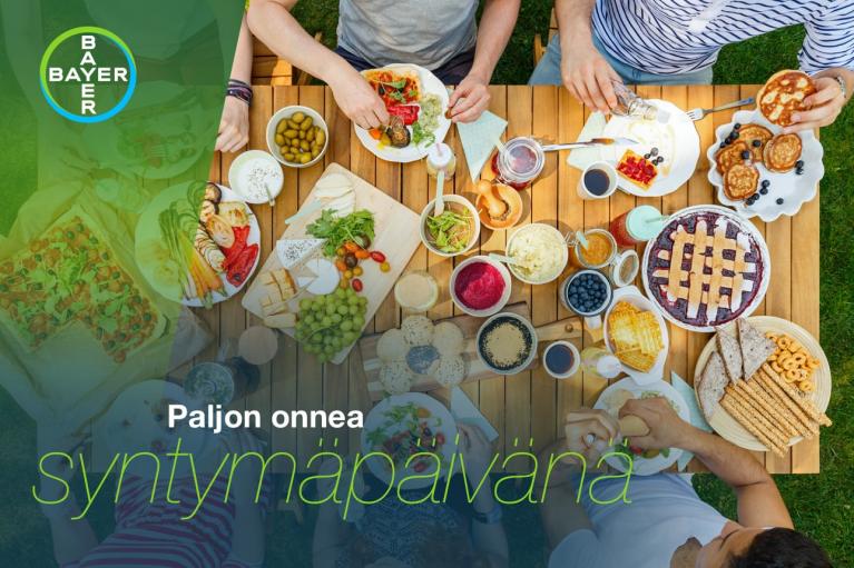 A group enjoys a meal at an outdoor table with various dishes, including pies and fruit. The Bayer logo is visible, and text reads "Paljon onnea syntymäpäivänä," meaning "Happy Birthday.