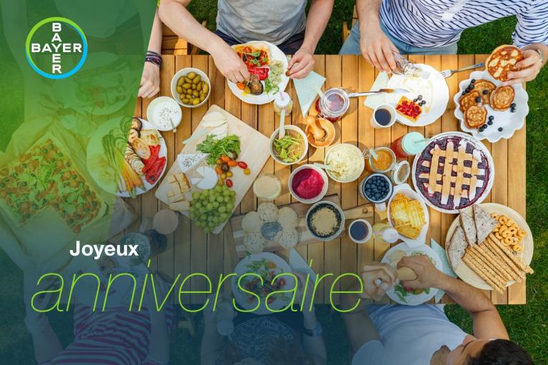 Top view of a table filled with various dishes, including pie, salad, and breadsticks, surrounded by people reaching for food. The text "Joyeux anniversaire" is overlaid in the image.