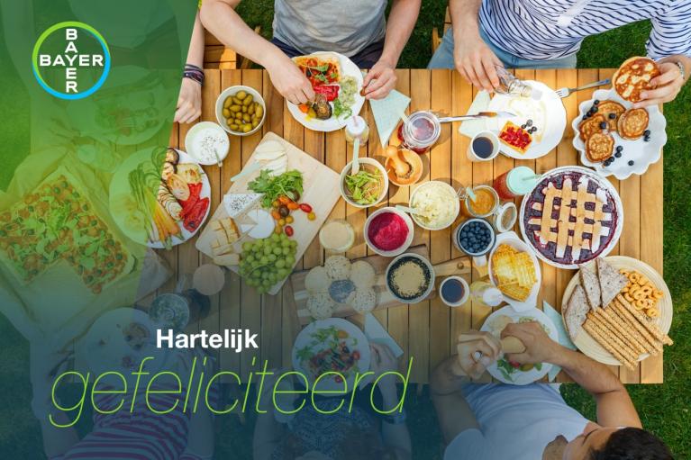 A top-down view of a group of people eating at an outdoor table laden with various foods. The image includes a "Bayer" logo and the text "Hartelijk gefeliciteerd.