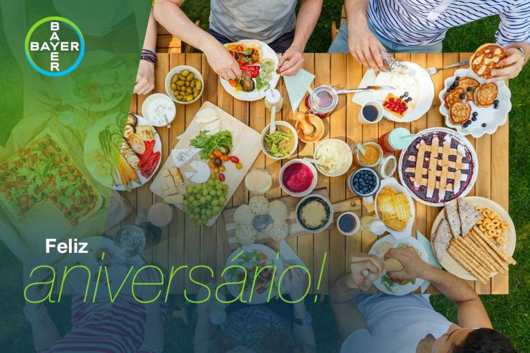Top-down view of a table with people enjoying a variety of foods and drinks outdoors, overlaid with a translucent Bayer logo and the text "Feliz aniversario!.