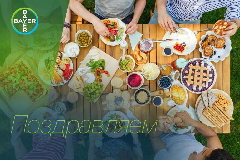 A table with a variety of dishes and people serving themselves; the Bayer logo is visible with "Поздравляем" text overlay.