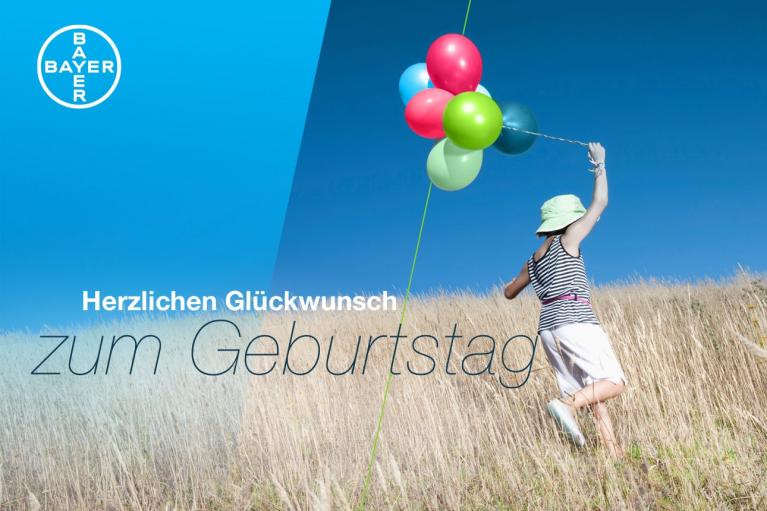 A person in a striped dress and hat runs through a field holding colorful balloons. The image includes the Bayer logo and the German text "Herzlichen Glückwunsch zum Geburtstag.