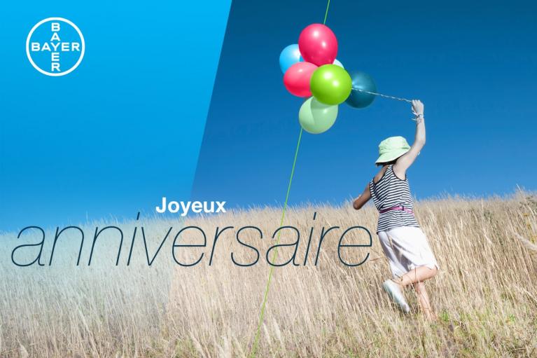 Person in a striped dress and hat runs through a field with colorful balloons. Text reads "Joyeux anniversaire." Bayer logo in the upper left corner.