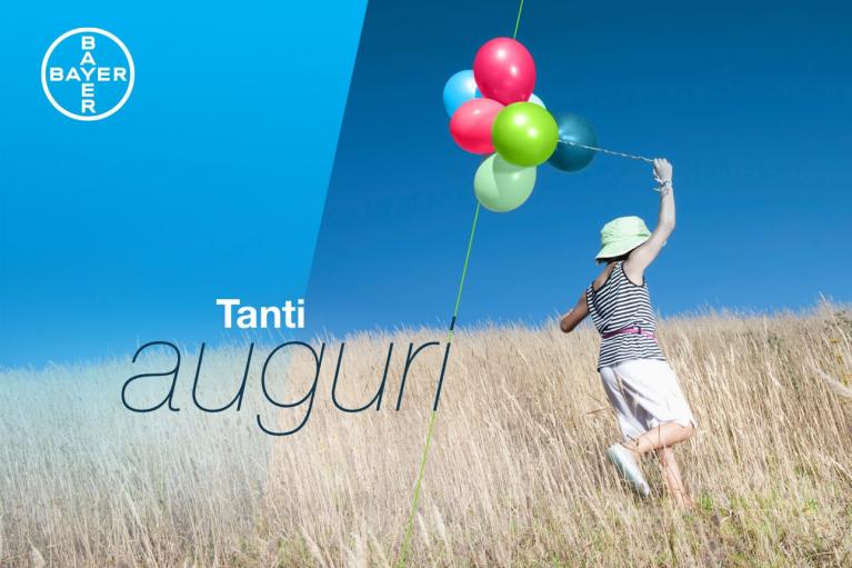 Child holding colorful balloons in a grassy field under a clear blue sky, with the words "Tanti auguri" and the Bayer logo.