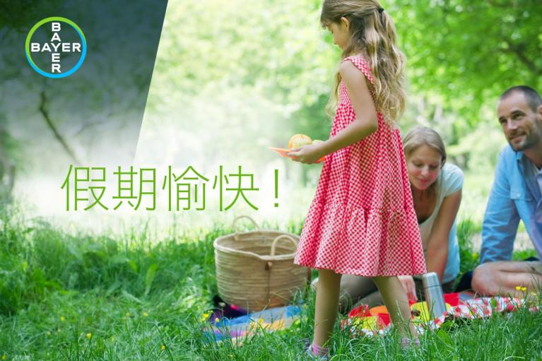 A girl in a red checkered dress stands in front of adults having a picnic on grass. The Bayer logo appears in the corner with Chinese text above.
