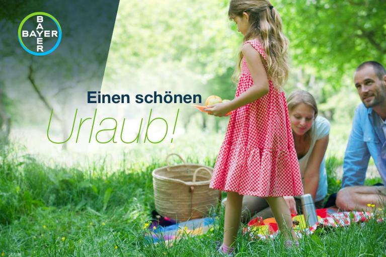 A family having a picnic outdoors on grass with a wicker basket. A child in a red dress holds an orange. "Einen schönen Urlaub!" text is displayed with the Bayer logo.