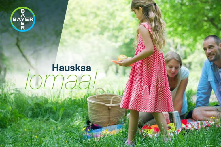 A family enjoys a picnic on grass with a wicker basket and food. A young girl in a red dress stands holding an orange. The words “Hauskaa lomaa!” and a Bayer logo are visible.