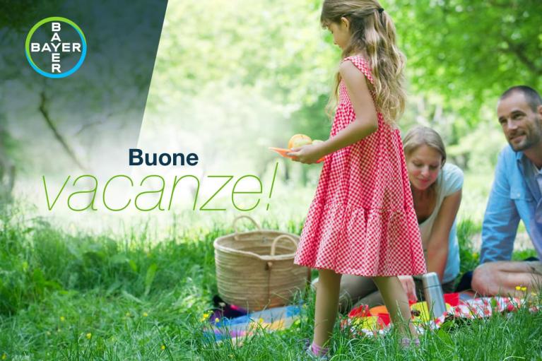 A family enjoys a picnic on the grass. A child in a red dress walks nearby. Text reads "Buone vacanze!" with a Bayer logo in the corner.