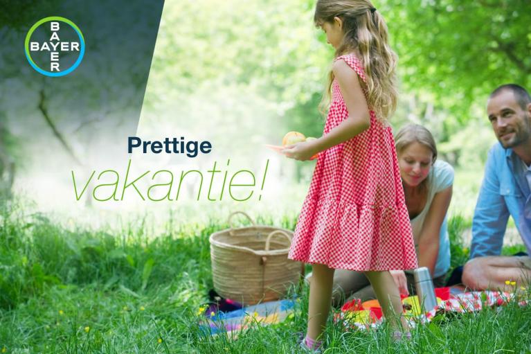 A family enjoys a picnic on a blanket in a grassy area. A child in a red dress stands while two adults sit. The text reads "Prettige vakantie!" with a Bayer logo.