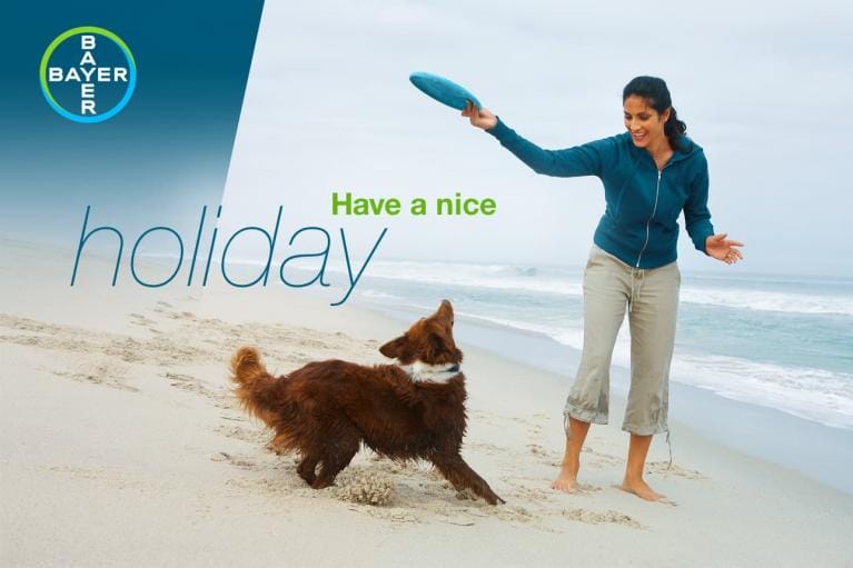 Woman playing fetch with a dog on a beach. Text reads "Have a nice holiday" next to the Bayer logo.