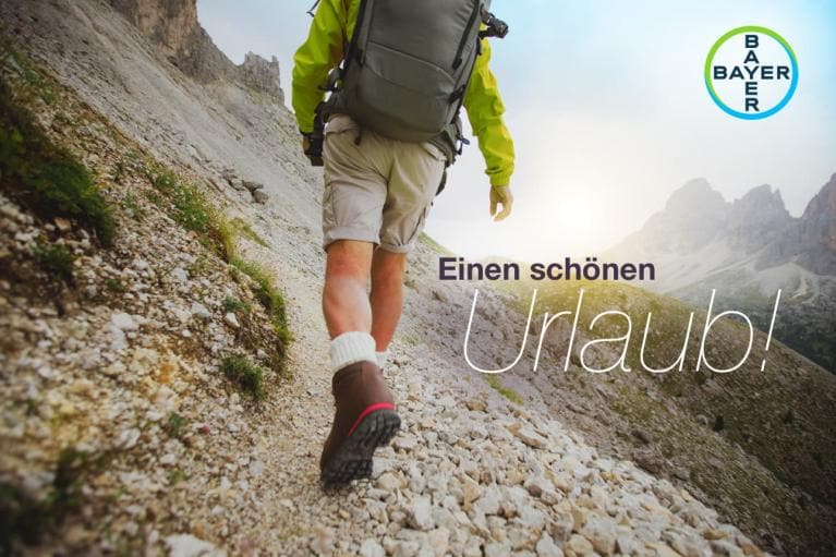A person hiking on a rocky trail with mountains in the background. The text reads "Einen schönen Urlaub!" and includes the Bayer logo.