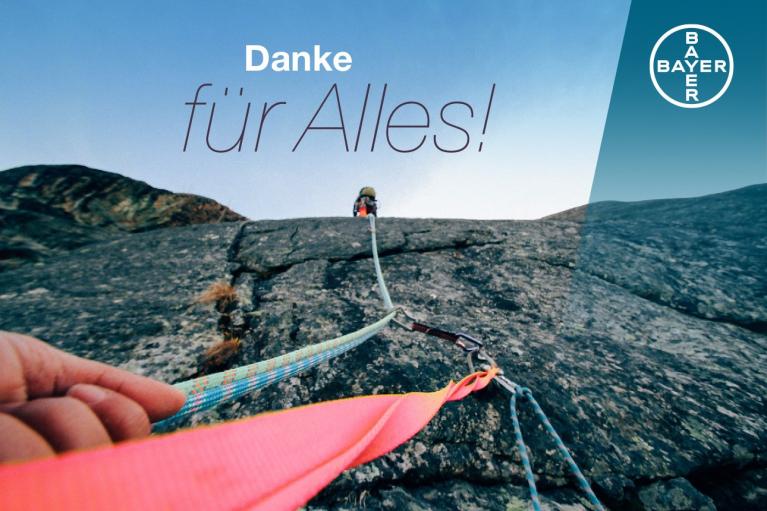 View from below of a person climbing a rock face, secured by ropes. Text reads "Danke für Alles!" and features the Bayer logo in the top right corner.