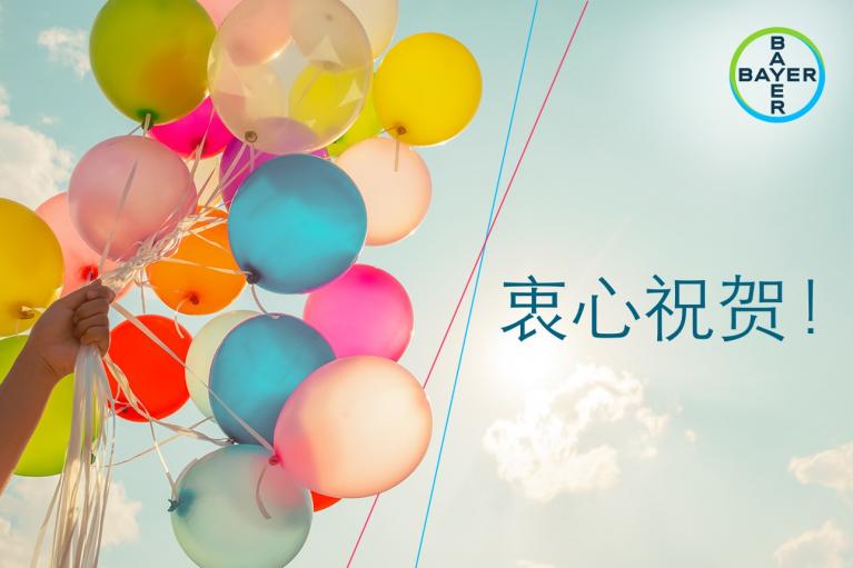 A hand holds colorful balloons against a blue sky. "衷心祝贺!" and the Bayer logo appear on the right.