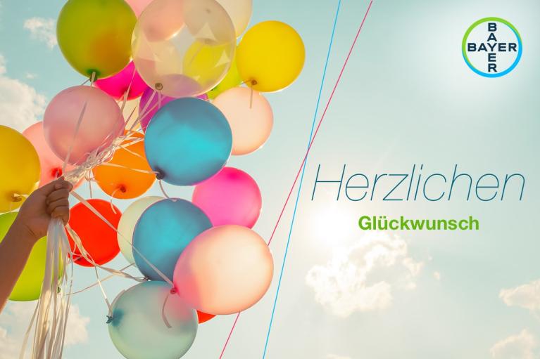 A hand holds colorful balloons against a sky background. Text reads "Herzlichen Glückwunsch" with a Bayer logo in the corner.