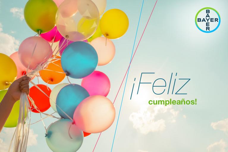 A hand holds colorful balloons against a blue sky. Text reads "¡Feliz cumpleaños!" with a Bayer logo in the top right.