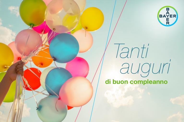 Colorful balloons held against a sky with clouds, accompanied by the text "Tanti auguri di buon compleanno" and a Bayer logo.