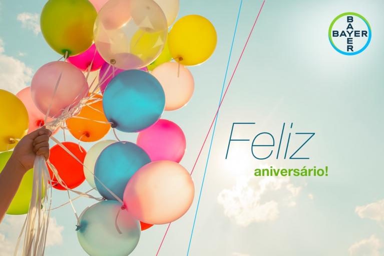 A hand holds colorful balloons in a bright sky. The text reads "Feliz aniversário!" with the Bayer logo in the top right corner.