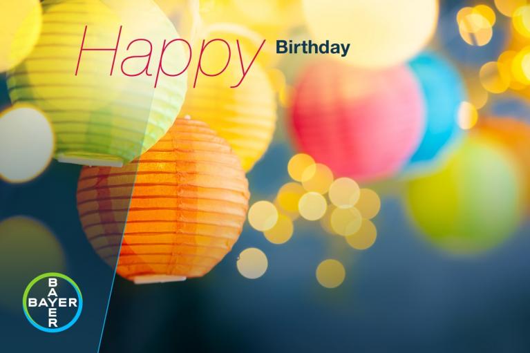 Colorful paper lanterns with the words "Happy Birthday" and a blurred background of yellow bokeh lights. Bayer logo in the corner.