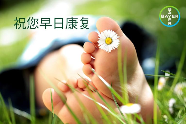 Close-up of bare feet resting in grass with a daisy between toes, Bayer logo in corner, Chinese text on the side.