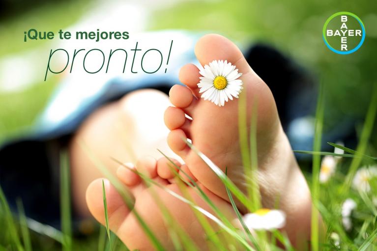 Close-up of bare feet in grass with a daisy between toes and "¡Que te mejores pronto!" text. Bayer logo in the top right corner.