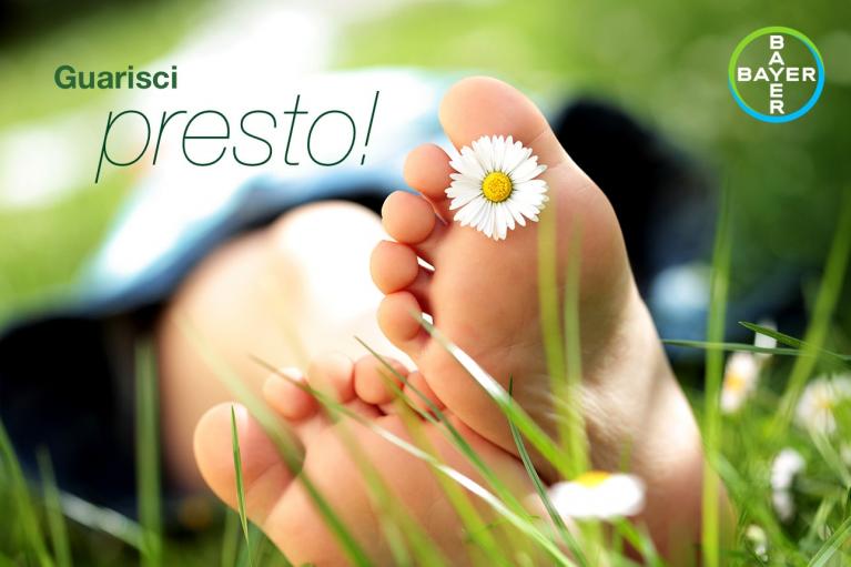 Close-up of bare feet with a daisy on toes, lying on grass. The text reads "Guarisci presto!" with a Bayer logo in the top right corner.