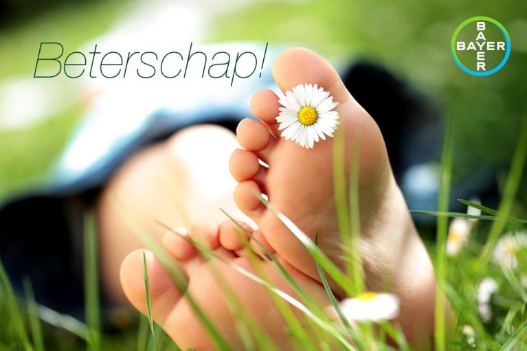 Close-up of a person's bare foot with a daisy resting on the toes. The word "Beterschap!" and a Bayer logo are visible.