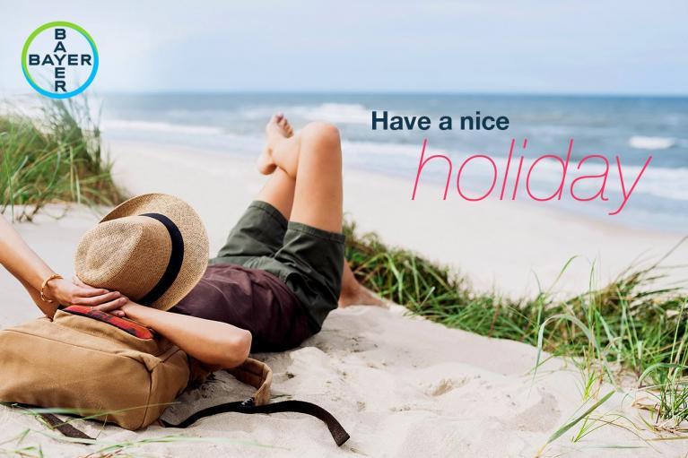 Person relaxing on a sandy beach with a straw hat and a backpack. Text reads, "Have a nice holiday." Bayer logo in the corner.