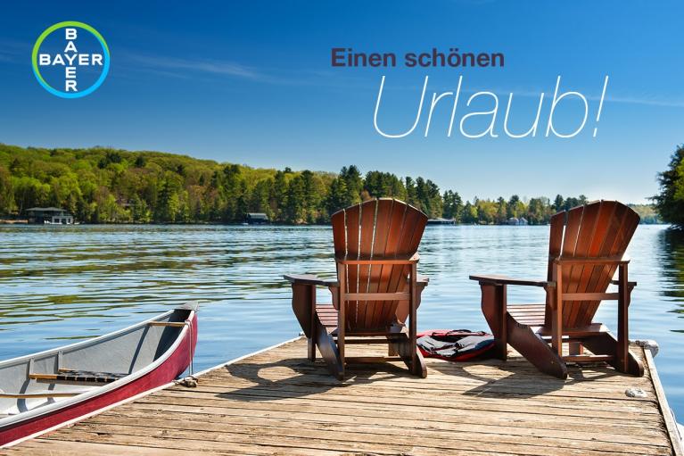 Two wooden chairs and a canoe on a dock facing a lake with forested shoreline. Text in German: "Einen schönen Urlaub!" Bayer logo in the corner.