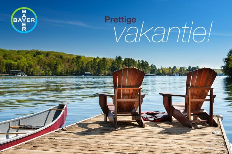 Two wooden chairs and a canoe on a dock by a lake under a clear blue sky. Trees in the background. Text reads "Prettige vakantie!" and a Bayer logo is visible.