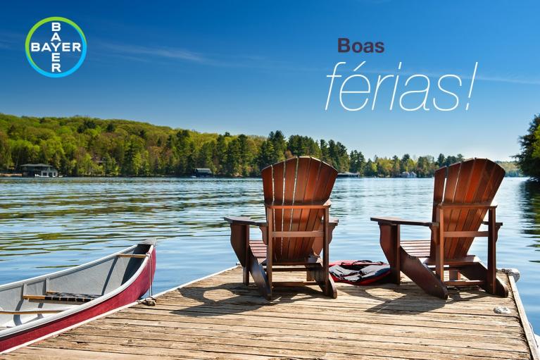 Two wooden chairs and a canoe on a dock overlooking a calm lake with a tree-lined shore, under a clear blue sky. Text reads "Boas férias!" and shows the Bayer logo.