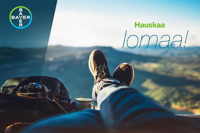 Person relaxing with legs extended, overlooking a scenic view. Bayer logo in top left corner. Finnish text reads "Hauskaa lomaa!" with camera and backpack nearby.