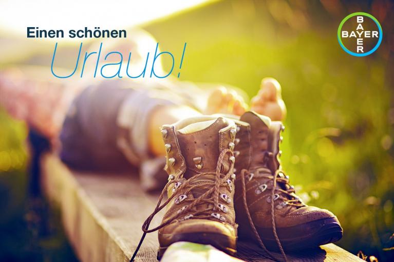 A person lying on a wooden bench with hiking boots in the foreground. The background is sunlit and blurred. Text reads "Einen schönen Urlaub!" with a Bayer logo in the corner.